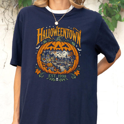 May include: A navy blue t-shirt featuring a large orange jack-o'-lantern graphic with a detailed town scene inside. The text reads HALLOWEENTOWN and EST. 1998, accompanied by small bats, stars, and autumn leaves surrounding the central pumpkin design.