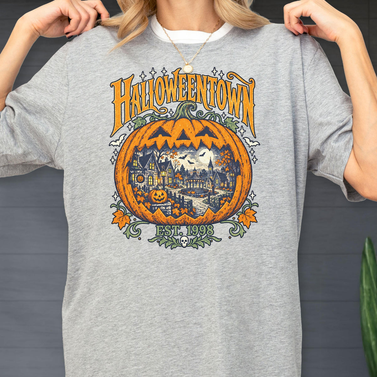 May include: A heather gray t-shirt featuring a large graphic of a carved jack-o'-lantern containing a spooky village scene. The text reads HALLOWEENTOWN and EST. 1998, accented with bats, stars, and autumn leaves in orange, yellow, and dark blue tones.