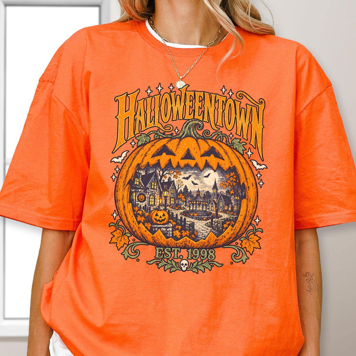 May include: An orange short-sleeve t-shirt featuring a large graphic of a jack-o'-lantern containing a spooky town scene. The text reads HALLOWEENTOWN EST. 1998. The design includes small bats, stars, and autumn leaves in shades of yellow, green, and black.
