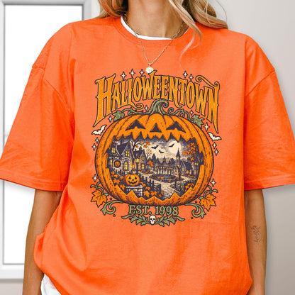 May include: An orange short-sleeve t-shirt featuring a large graphic of a jack-o'-lantern containing a spooky town scene. The text reads HALLOWEENTOWN EST. 1998. The design includes small bats, stars, and autumn leaves in shades of yellow, green, and black.