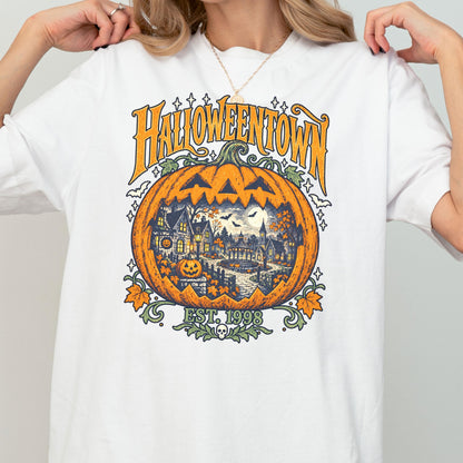 May include: A white t-shirt featuring a large orange pumpkin graphic with a detailed scene of a spooky town inside. The text reads HALLOWEENTOWN and EST. 1998. The design includes small bats, stars, and a skull icon, rendered in orange, green, and dark blue.
