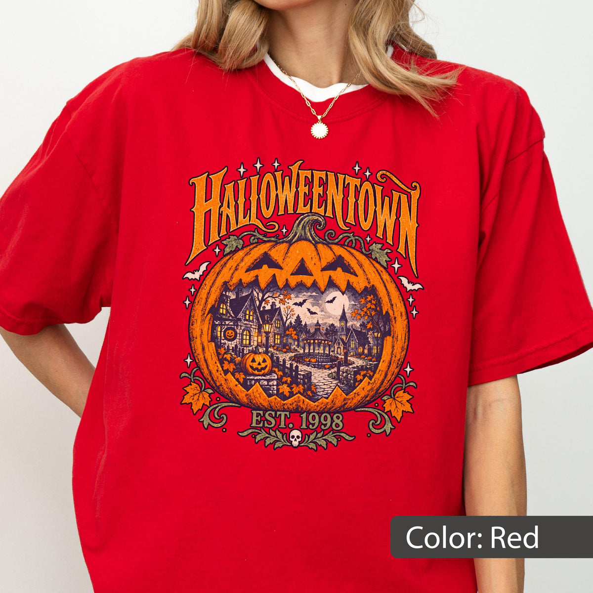 May include: A red t-shirt featuring a large orange pumpkin graphic with the text HALLOWEENTOWN and EST. 1998. The pumpkin contains a detailed illustration of a spooky village scene with houses, trees, bats, and a full moon, accented by small stars.
