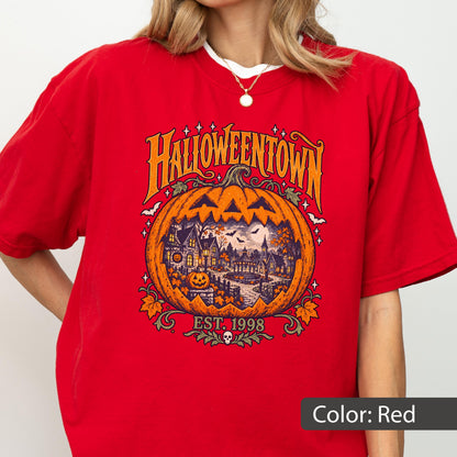 May include: A red t-shirt featuring a large orange pumpkin graphic with the text HALLOWEENTOWN and EST. 1998. The pumpkin contains a detailed illustration of a spooky village scene with houses, trees, bats, and a full moon, accented by small stars.