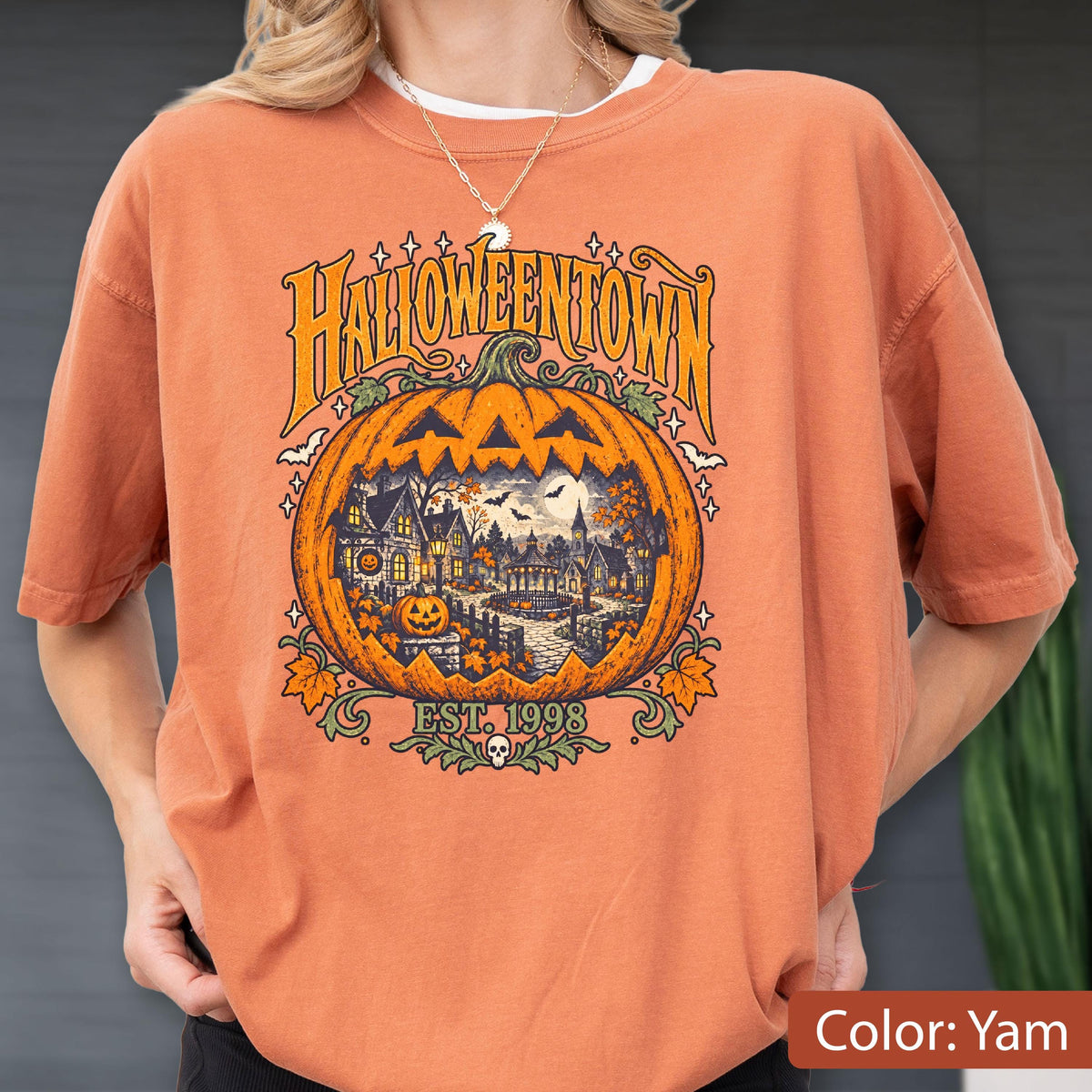 May include: A burnt orange t-shirt featuring a large graphic of a carved jack-o'-lantern pumpkin. Inside the pumpkin is a detailed scene of a spooky town with houses, trees, and bats. The text reads HALLOWEENTOWN and EST. 1998.