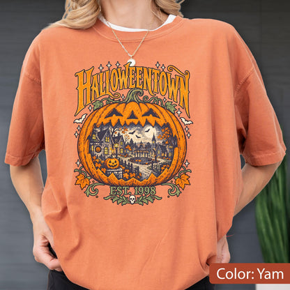 May include: A burnt orange t-shirt featuring a large graphic of a carved jack-o'-lantern pumpkin. Inside the pumpkin is a detailed scene of a spooky town with houses, trees, and bats. The text reads HALLOWEENTOWN and EST. 1998.