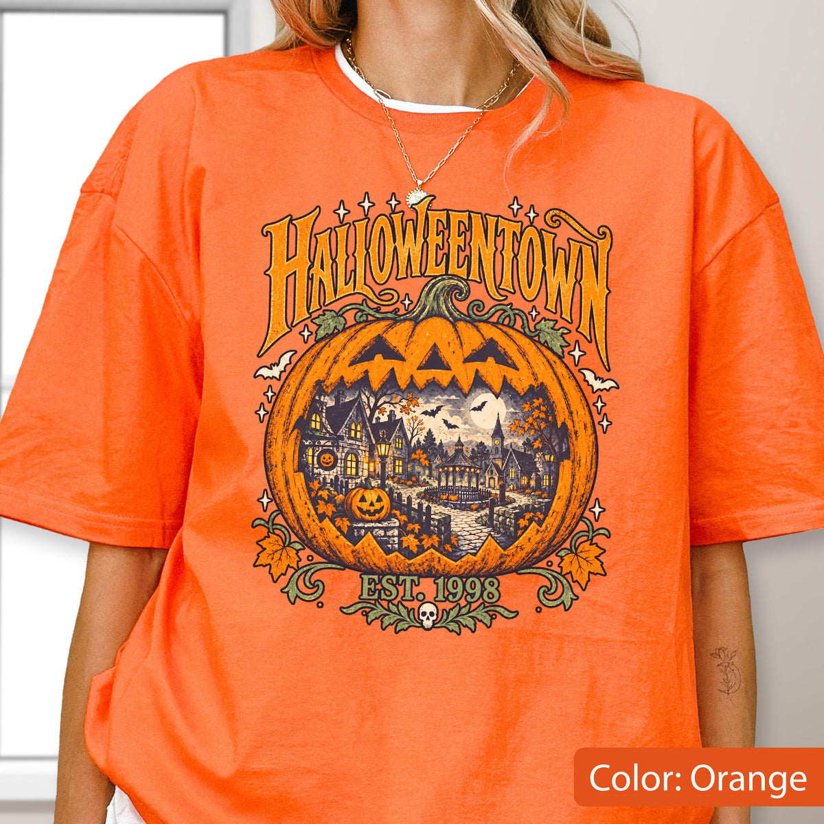 May include: An orange t-shirt featuring a large graphic of a carved pumpkin with a detailed scene of a town inside. The text reads HALLOWEENTOWN and EST. 1998. The design includes small bats, stars, and a skull icon at the bottom, rendered in shades of yellow, brown, and green.