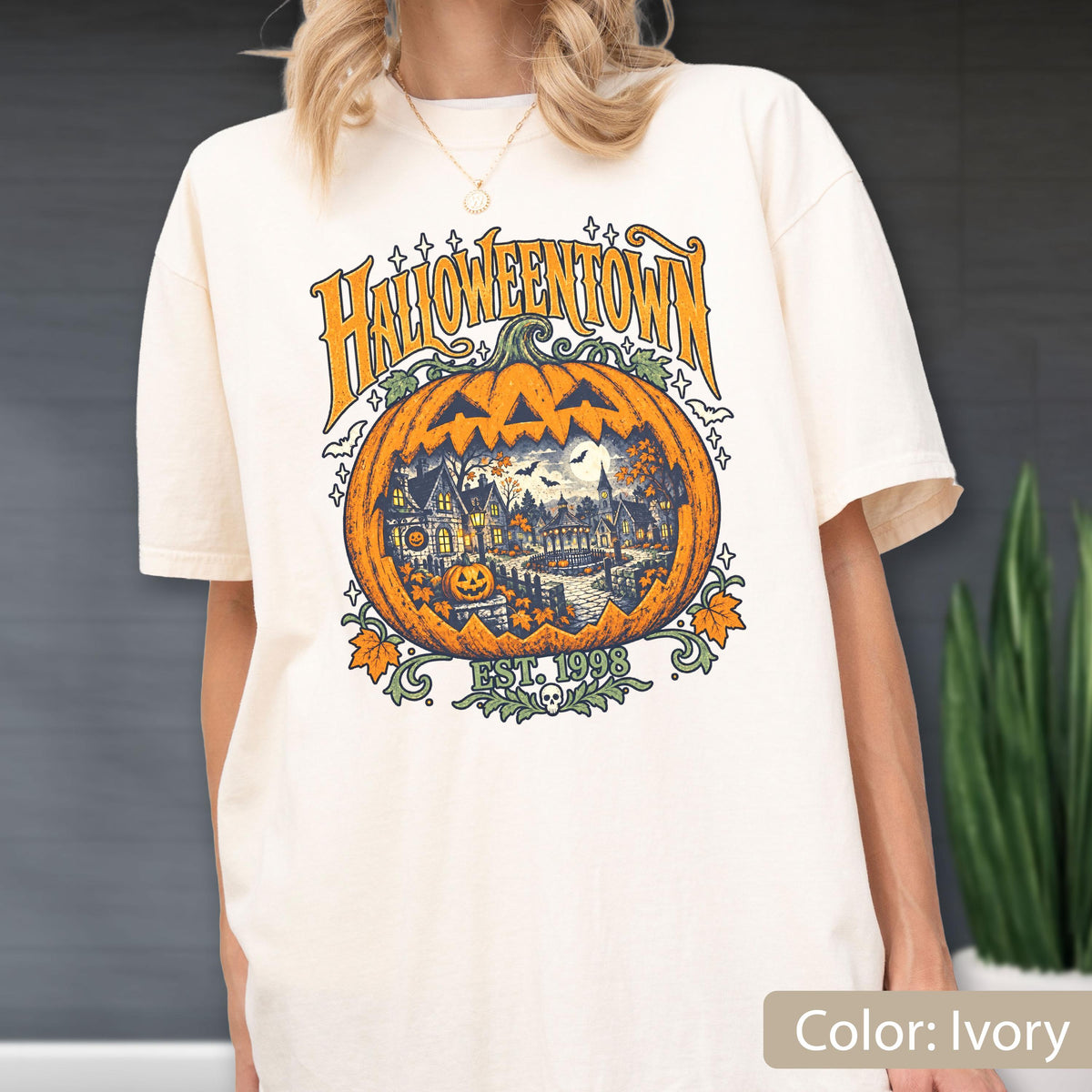 May include: An ivory t-shirt featuring a large graphic of a carved pumpkin with a town scene inside. The text reads HALLOWEENTOWN and EST. 1998. The design includes orange, yellow, and green colors with small bats, stars, and a skull icon.
