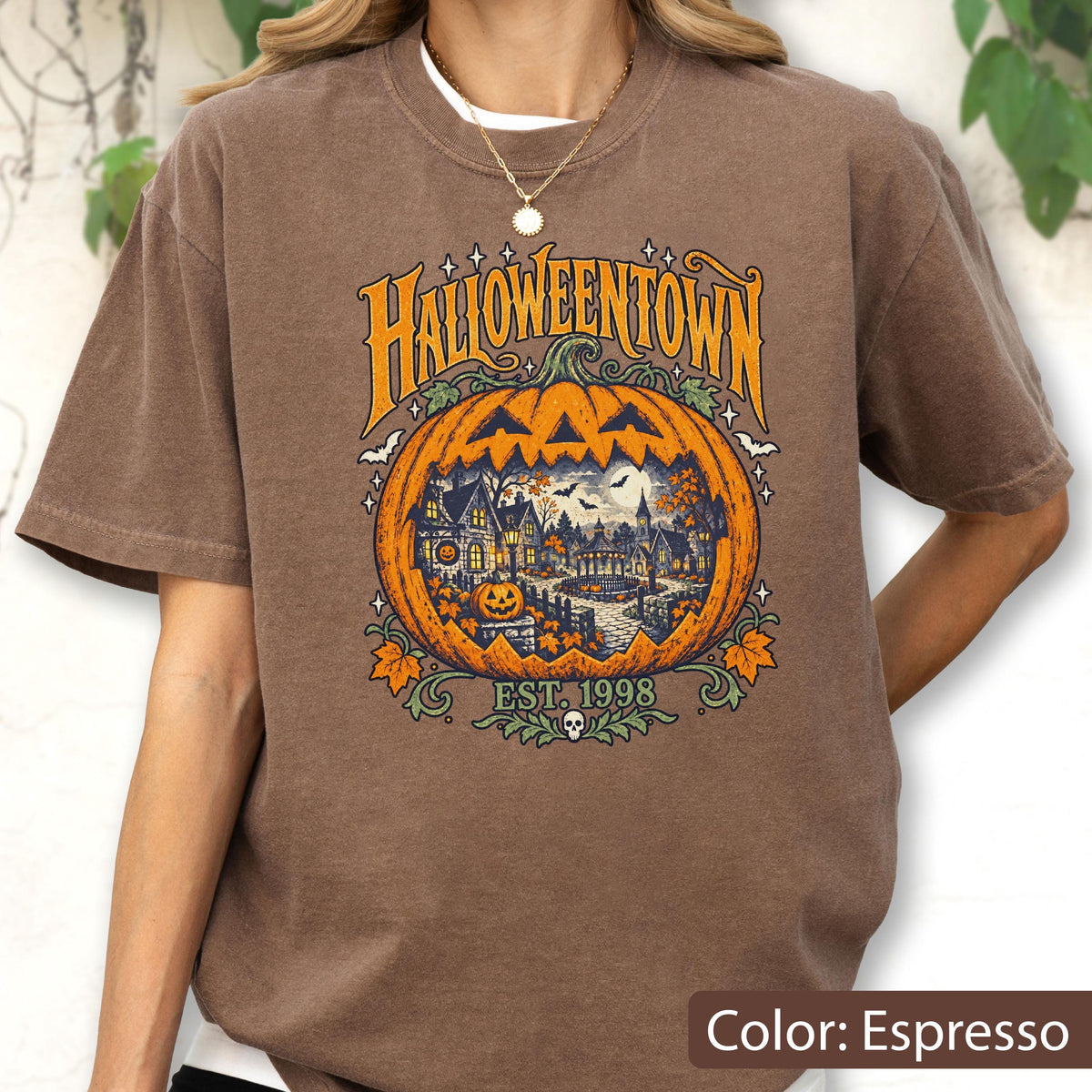 May include: An espresso brown t-shirt featuring a large graphic of a carved pumpkin with a spooky town scene inside. The text reads HALLOWEENTOWN and EST. 1998. The design includes orange, yellow, and dark blue colors with small stars and leaves.