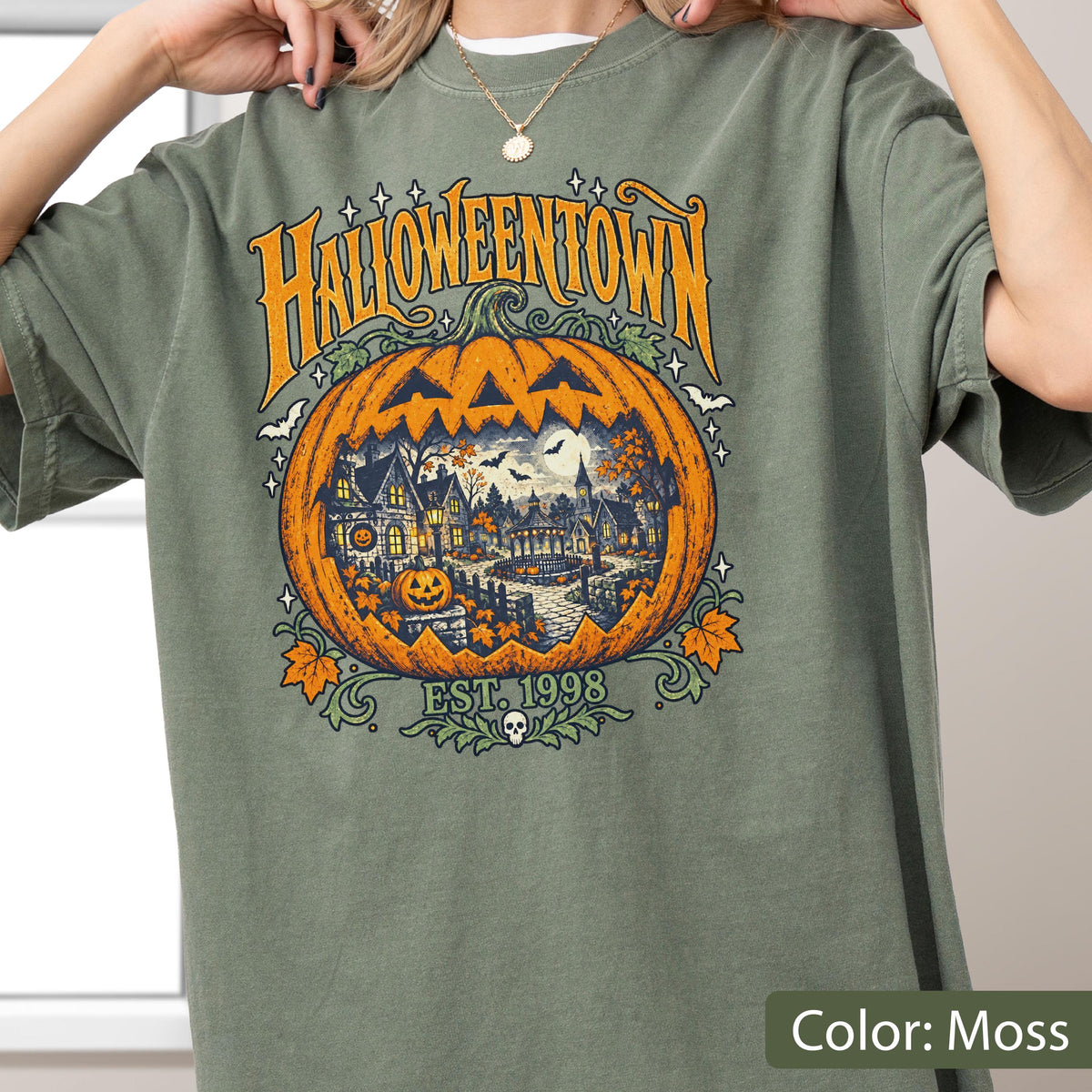 May include: A moss green t-shirt featuring a large graphic print of a carved orange pumpkin. Inside the pumpkin is a detailed scene of a town with houses, trees, and a full moon. The text reads HALLOWEENTOWN and EST. 1998 with a small skull icon.