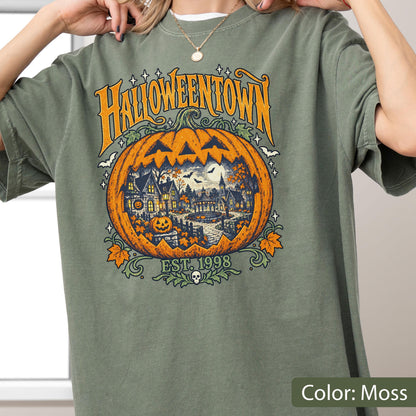 May include: A moss green t-shirt featuring a large graphic print of a carved orange pumpkin. Inside the pumpkin is a detailed scene of a town with houses, trees, and a full moon. The text reads HALLOWEENTOWN and EST. 1998 with a small skull icon.