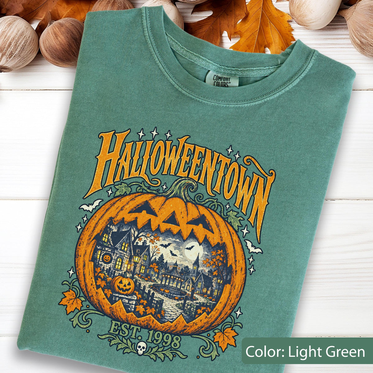 May include: A light green t-shirt featuring a graphic of a large orange jack-o'-lantern with a detailed scene of a town inside. The text reads HALLOWEENTOWN and EST. 1998. The design includes small bats, stars, and autumn leaves surrounding the pumpkin.
