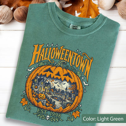 May include: A light green t-shirt featuring a graphic of a large orange jack-o'-lantern with a detailed scene of a town inside. The text reads HALLOWEENTOWN and EST. 1998. The design includes small bats, stars, and autumn leaves surrounding the pumpkin.
