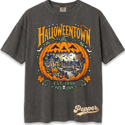 May include: A charcoal gray short-sleeve t-shirt featuring a large orange jack-o'-lantern graphic with a detailed town scene inside. The text reads HALLOWEENTOWN, EST. 1998, and Pepper. The design includes bats, stars, and autumn leaves.