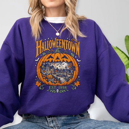 May include: A purple crewneck sweatshirt featuring a large orange jack-o'-lantern graphic with a detailed town scene inside. The text reads HALLOWEENTOWN and EST. 1998, accented with small stars, bats, and autumn leaves in orange, yellow, and green tones.