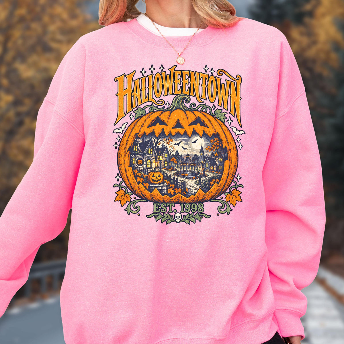 May include: A bright pink crewneck sweatshirt featuring a large graphic of a carved orange pumpkin. Inside the pumpkin is a detailed scene of a spooky town with houses, trees, and bats. The text reads HALLOWEENTOWN and EST. 1998.