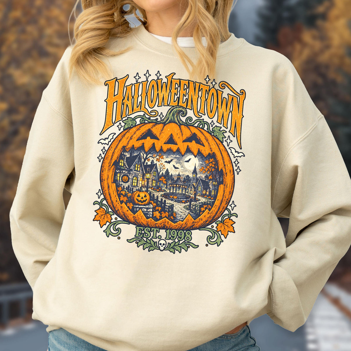 May include: A beige crewneck sweatshirt featuring a large graphic print of a carved orange pumpkin. Inside the pumpkin is a detailed scene of a spooky town with houses, trees, and a full moon. The text reads HALLOWEENTOWN and EST. 1998.