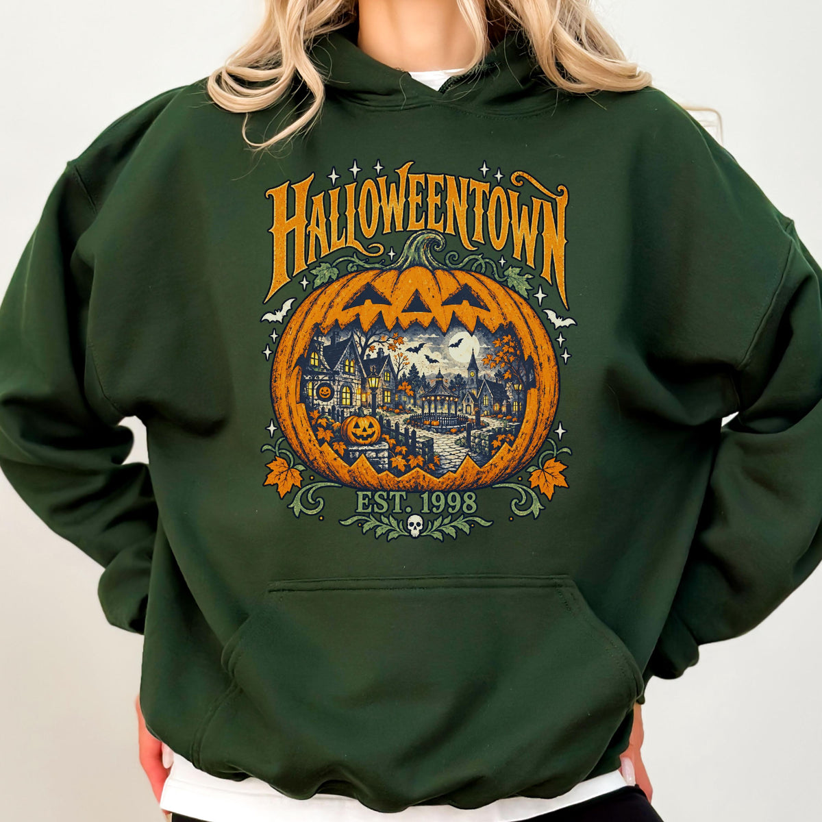 May include: A dark forest green hooded sweatshirt featuring a large graphic of a carved orange pumpkin. Inside the pumpkin is a detailed scene of a town with houses, trees, and a gazebo. The text reads HALLOWEENTOWN and EST. 1998 with a small skull.