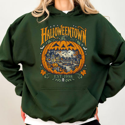 May include: A dark forest green hooded sweatshirt featuring a large graphic of a carved orange pumpkin. Inside the pumpkin is a detailed scene of a town with houses, trees, and a gazebo. The text reads HALLOWEENTOWN and EST. 1998 with a small skull.