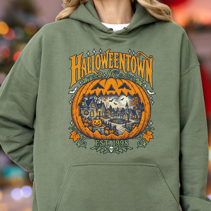May include: An olive green hooded sweatshirt featuring a graphic print of a large orange jack-o'-lantern. Inside the pumpkin is a detailed scene of a town with buildings, trees, and bats. The text reads HALLOWEENTOWN and EST. 1998.