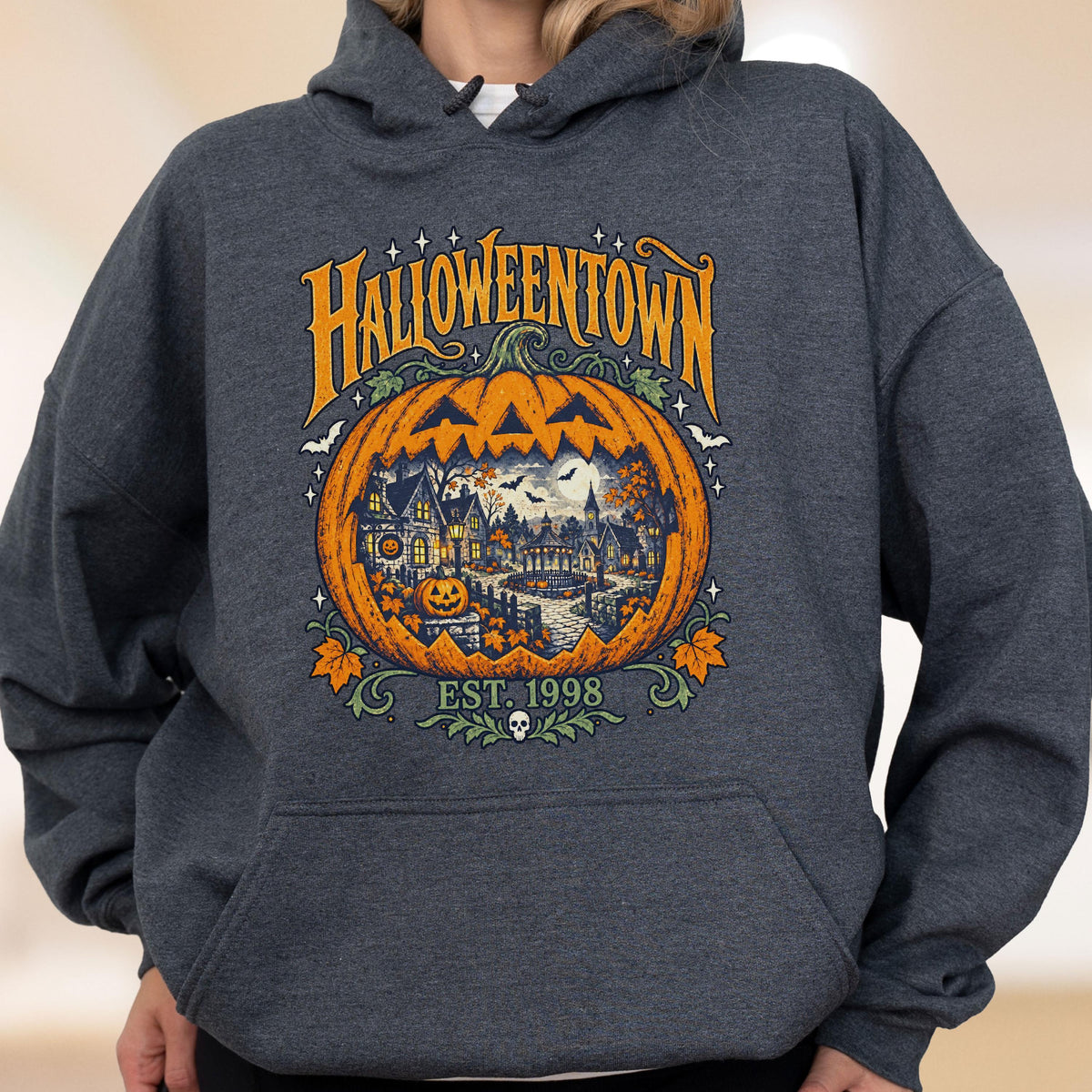 May include: A dark heather gray hooded sweatshirt featuring a large graphic print of a carved pumpkin. Inside the pumpkin is a detailed scene of a town with houses and trees. The text reads HALLOWEENTOWN and EST. 1998 with a small skull icon.
