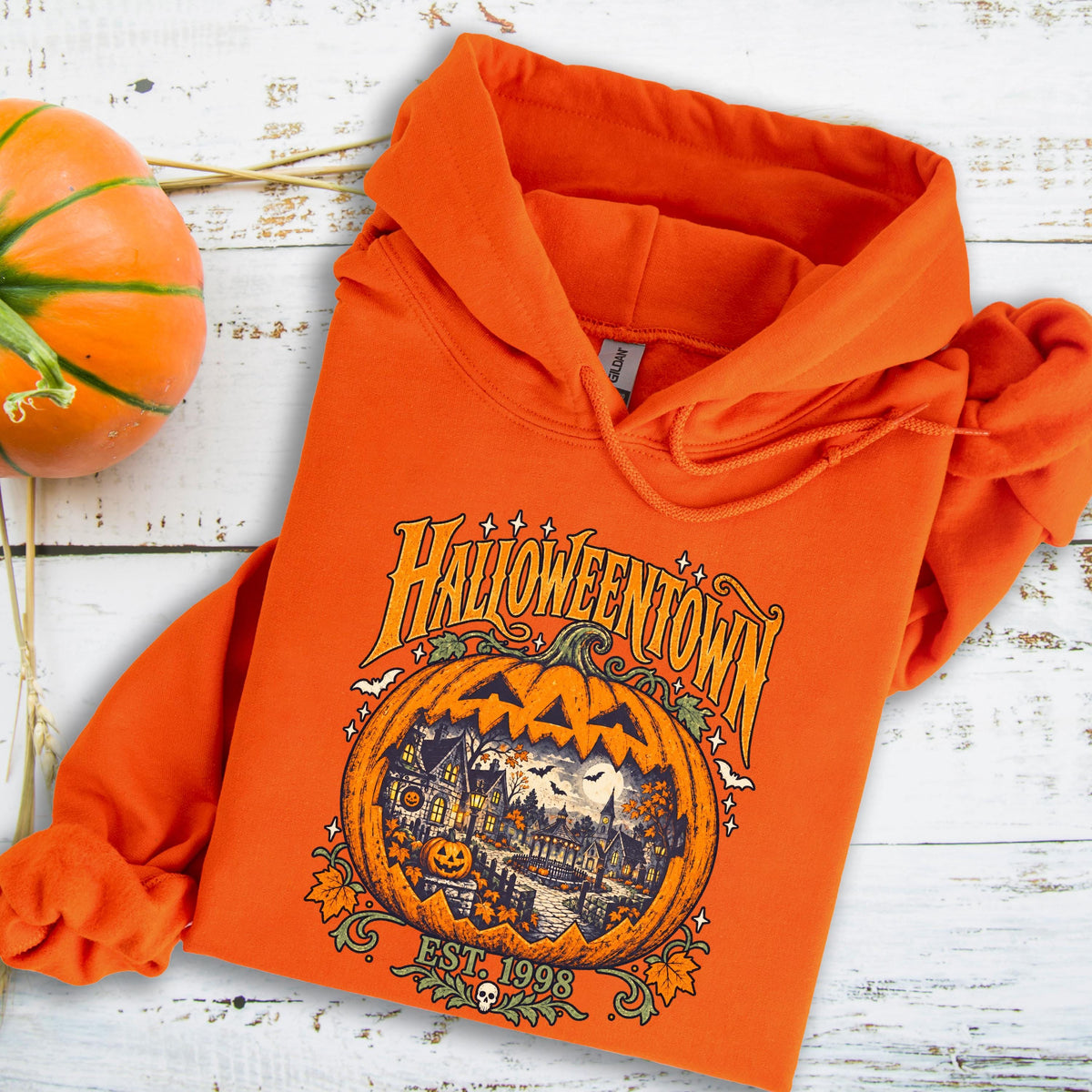 May include: An orange hooded sweatshirt featuring a graphic print of a carved pumpkin with a spooky house scene inside. The text HALLOWEENTOWN EST. 1998 is printed above and below the pumpkin. The hoodie has a drawstring hood and long sleeves.