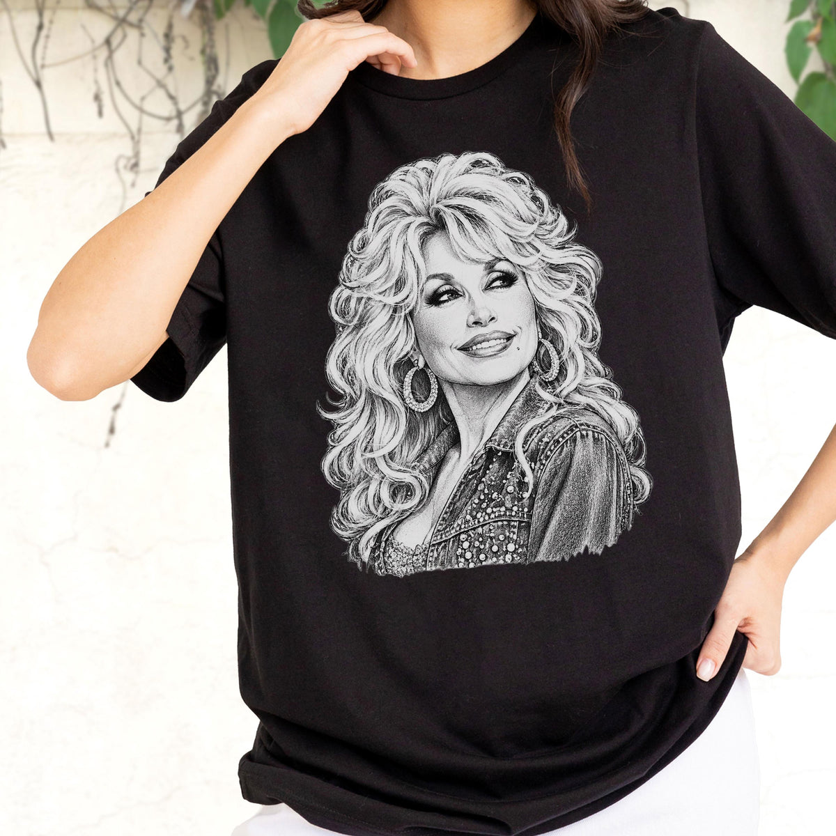 May include: A black short-sleeved t-shirt featuring a detailed white pencil-style sketch graphic of a person with voluminous blonde hair, hoop earrings, and a denim jacket. The illustration is centered on the chest of the garment.