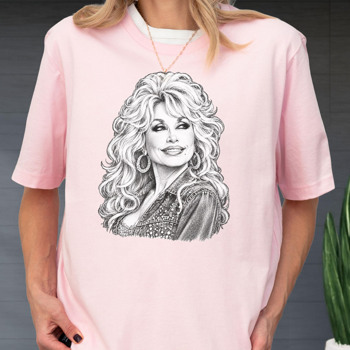 May include: A light pink short-sleeved t-shirt featuring a detailed black and white pencil-style sketch of a person with voluminous blonde hair, hoop earrings, and a denim jacket. The graphic is centered on the chest of the garment.