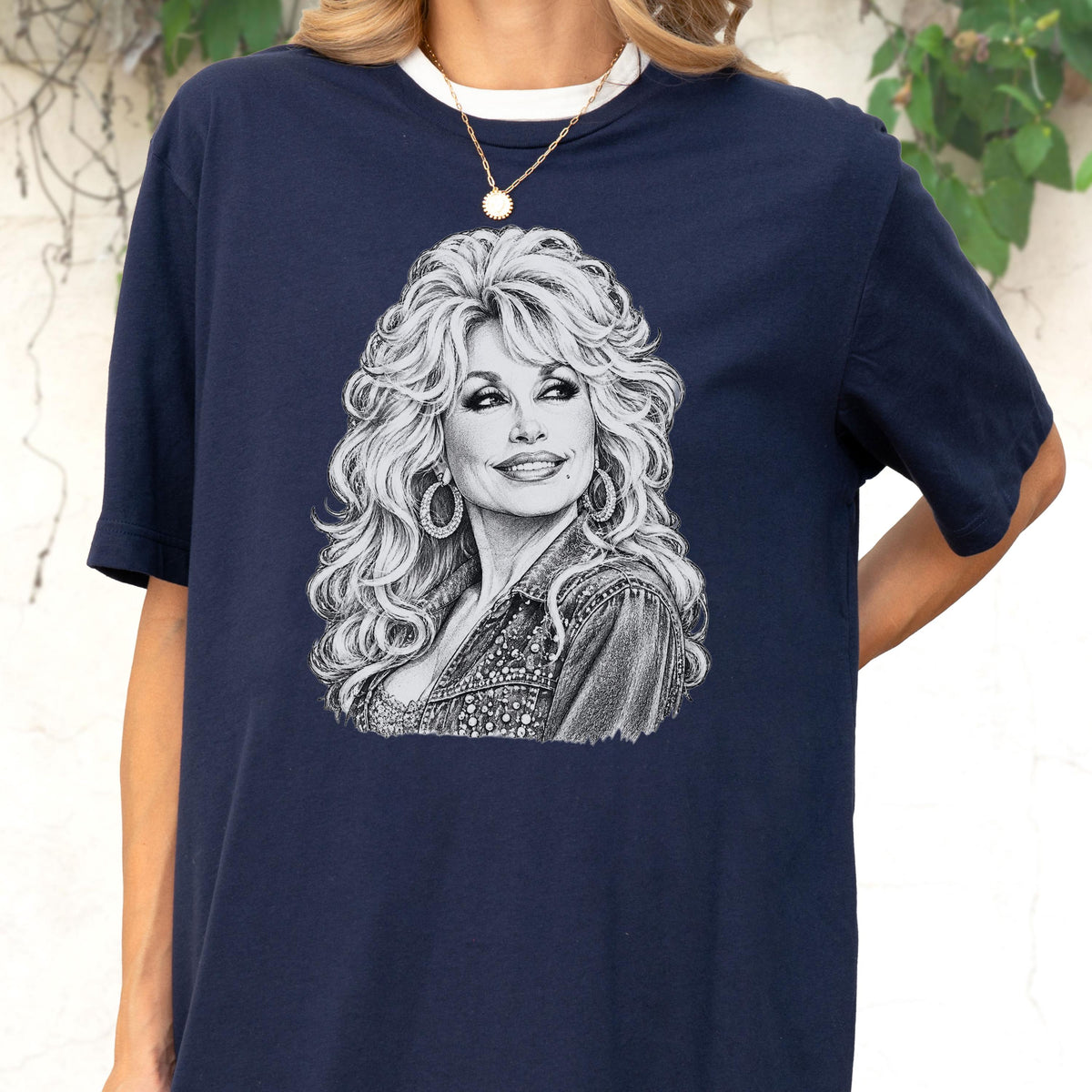 May include: A navy blue short-sleeved t-shirt featuring a detailed black and white graphic illustration of a person with voluminous blonde hair, large hoop earrings, and a denim jacket. The design is centered on the chest of the garment.