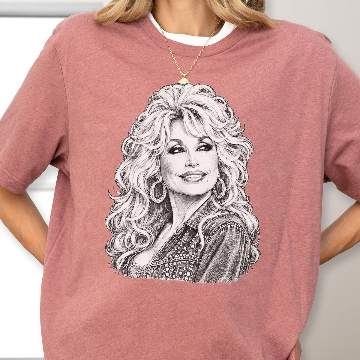 May include: A dusty rose short-sleeved t-shirt featuring a detailed black and white pencil-style sketch of a woman with voluminous blonde hair, hoop earrings, and a denim jacket. The graphic is centered on the chest of the crew neck garment.