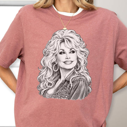 May include: A dusty rose short-sleeved t-shirt featuring a detailed black and white pencil-style sketch of a woman with voluminous blonde hair, hoop earrings, and a denim jacket. The graphic is centered on the chest of the crew neck garment.