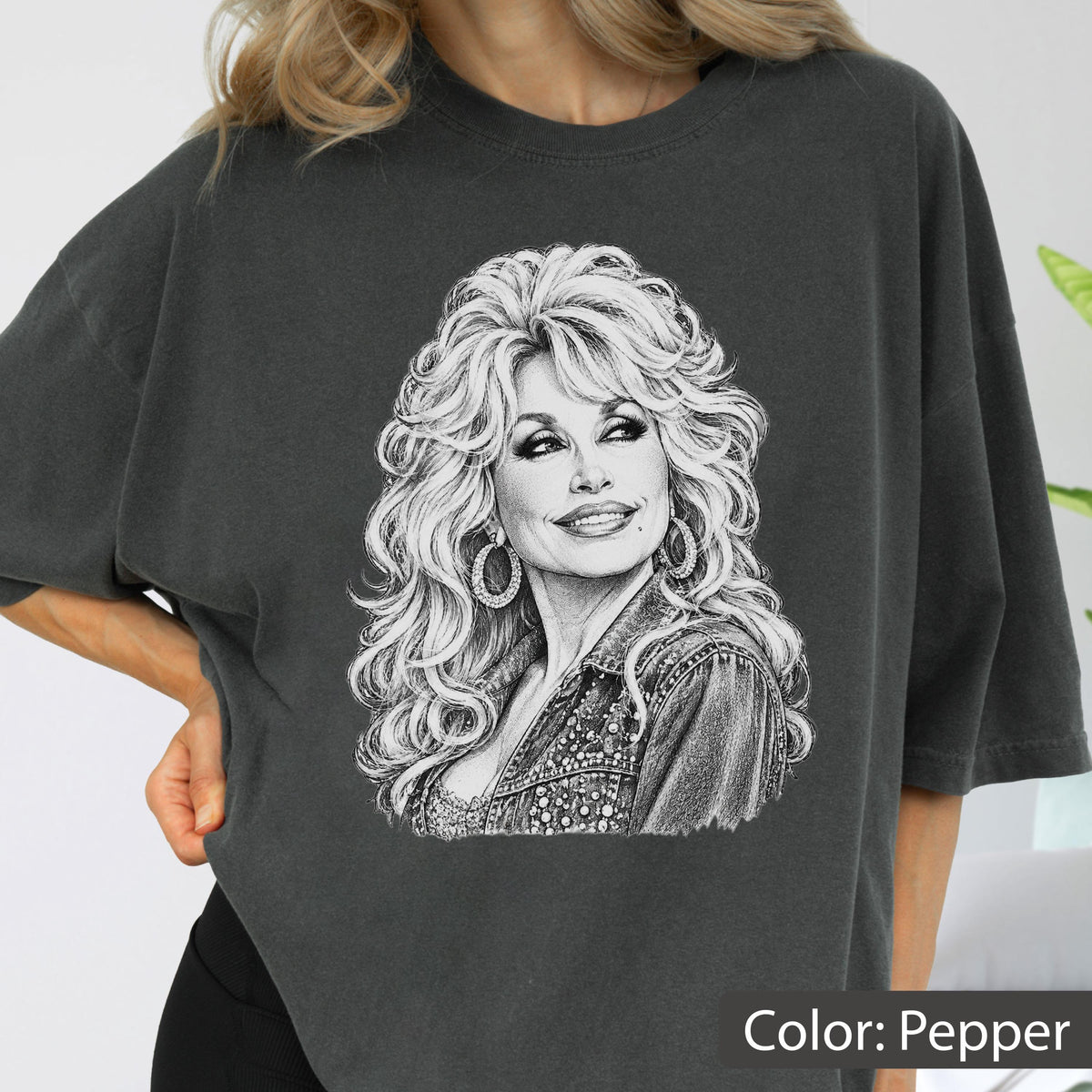 May include: A charcoal gray t-shirt featuring a detailed white line art illustration of a person with voluminous wavy hair, wearing hoop earrings and a textured jacket. The graphic is centered on the chest of the short-sleeved crew neck garment.