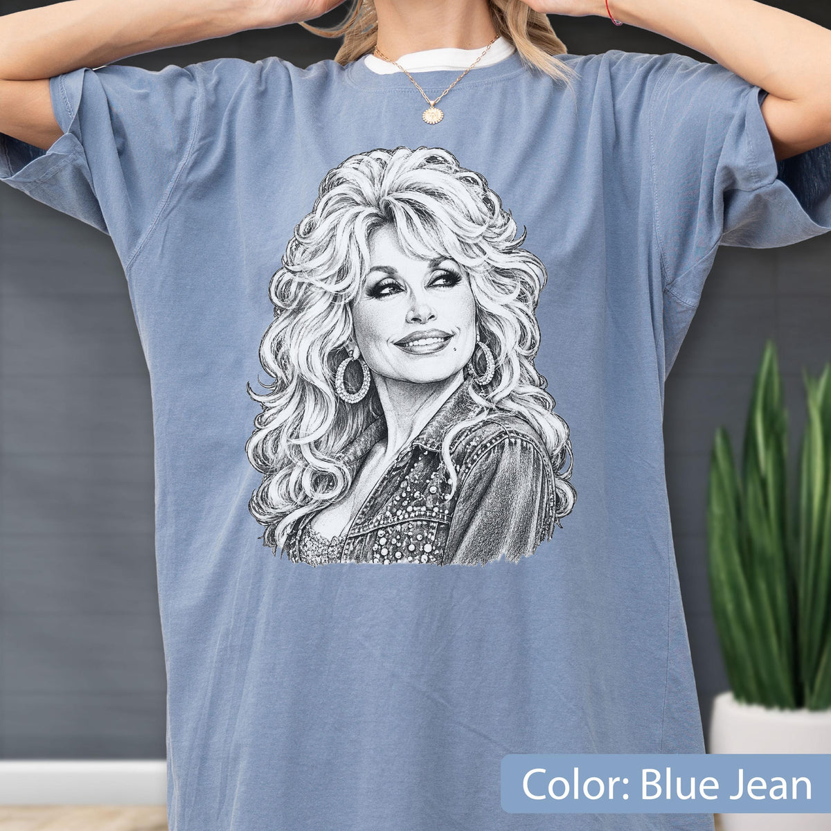 May include: A blue jean colored t-shirt featuring a detailed black and white sketch illustration of a woman with voluminous blonde hair, wearing hoop earrings and a textured jacket. The graphic is centered on the chest of the short-sleeved garment.