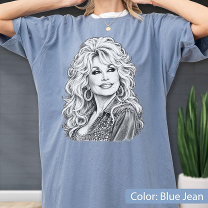 May include: A blue jean colored t-shirt featuring a detailed black and white sketch illustration of a woman with voluminous blonde hair, wearing hoop earrings and a textured jacket. The graphic is centered on the chest of the short-sleeved garment.