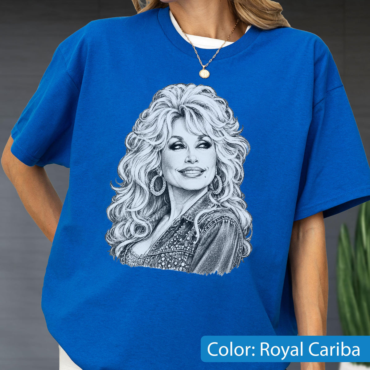 May include: A royal blue short-sleeved t-shirt featuring a detailed black and white sketch-style graphic of a person with voluminous hair, hoop earrings, and a textured jacket. The shirt has a crew neckline and a relaxed, boxy fit.