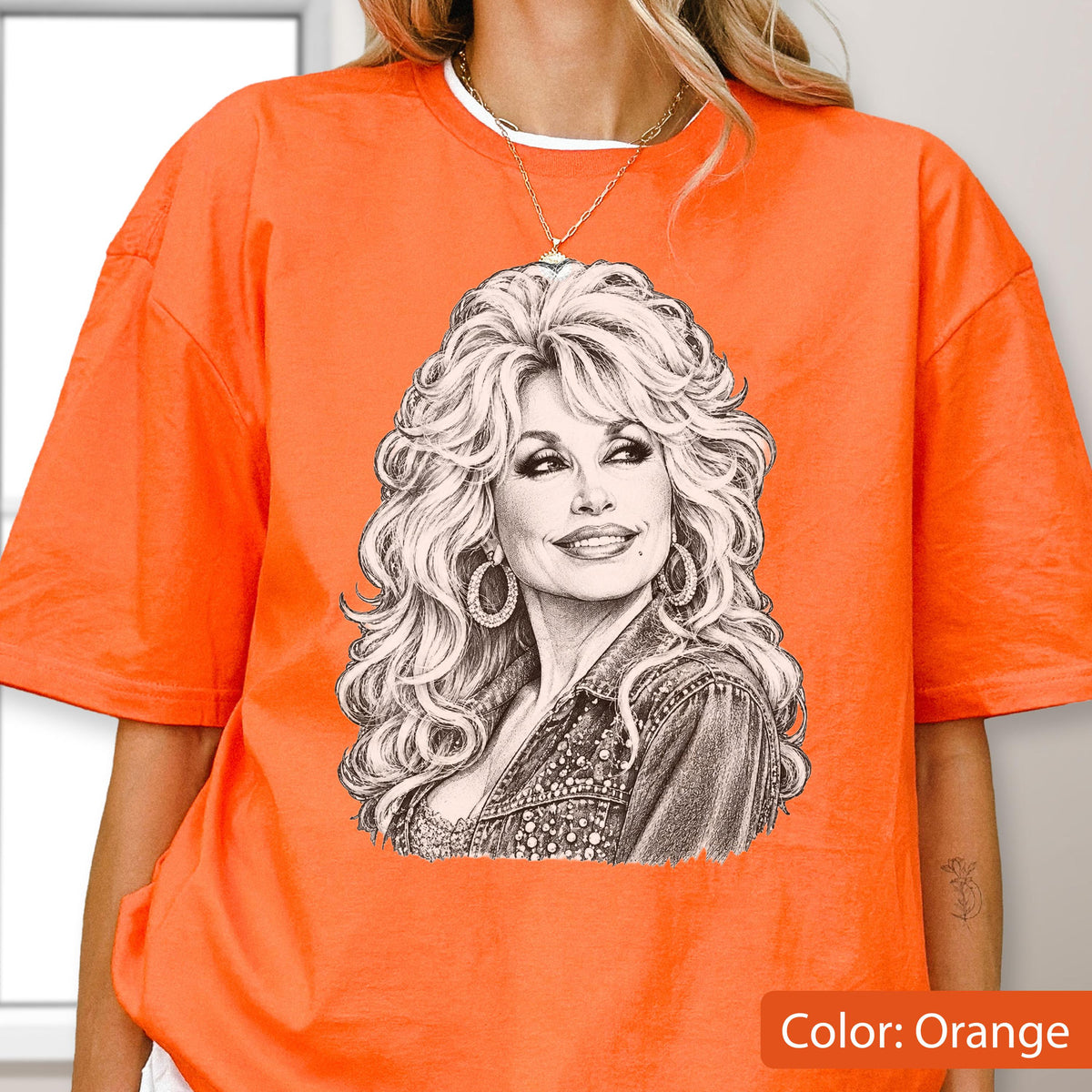 May include: A bright orange short-sleeved t-shirt featuring a detailed black and white pencil-style sketch of a woman with voluminous blonde hair, hoop earrings, and a denim jacket. The graphic is centered on the chest of the garment.