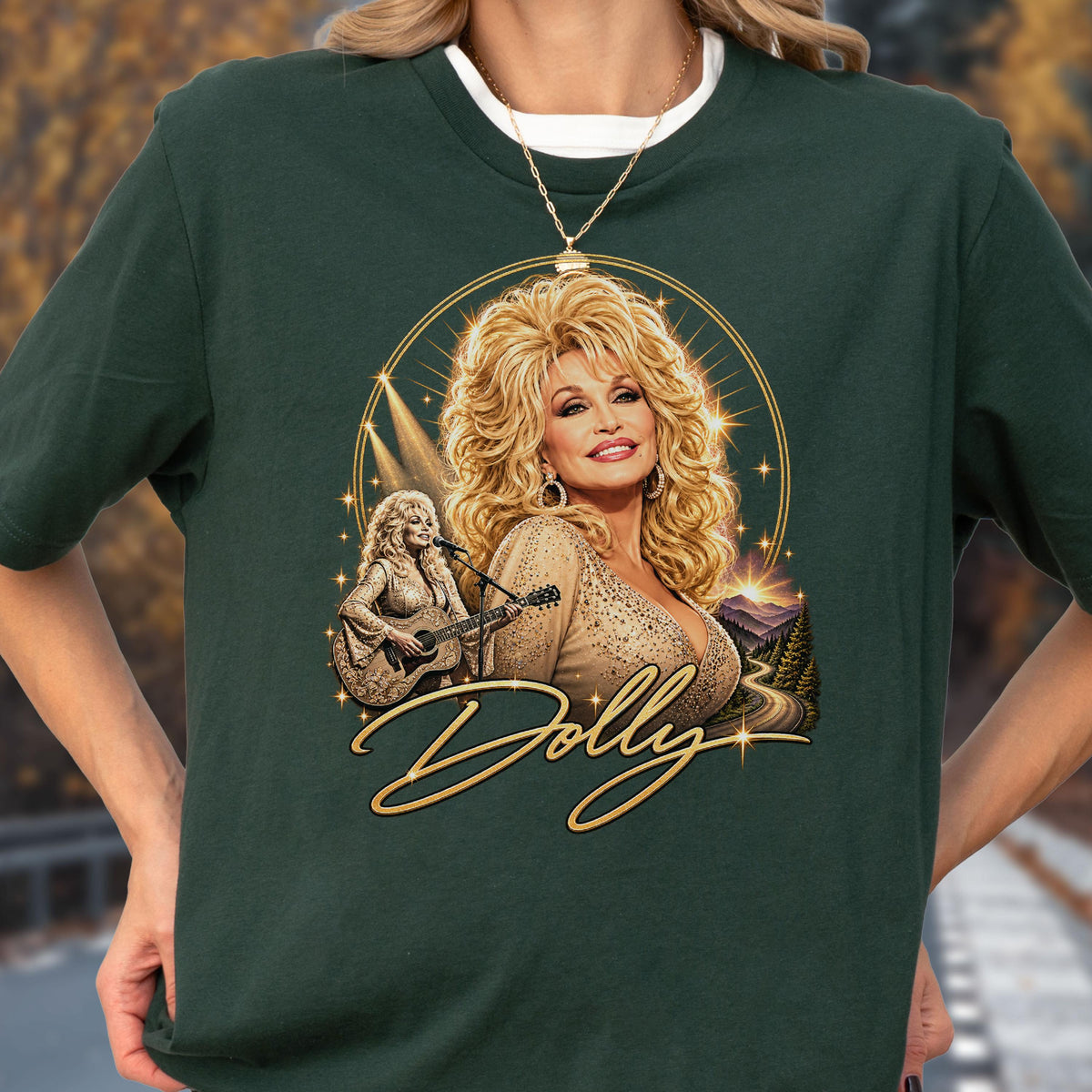 May include: A dark green t-shirt featuring a graphic print of Dolly Parton with blonde hair, wearing a beige top. The design includes a smaller image of her playing an acoustic guitar, a mountain landscape, and the word Dolly in gold script lettering.