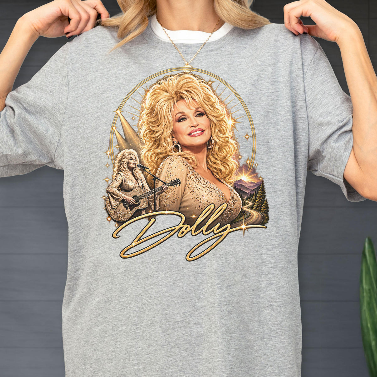 May include: A heather gray t-shirt featuring a graphic print of a blonde woman with voluminous hair, wearing a shimmering top. The design includes a smaller image of the woman playing an acoustic guitar, a mountain landscape, and the script text Dolly.