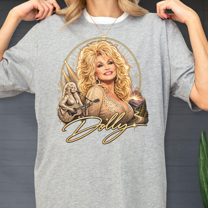 May include: A heather gray t-shirt featuring a graphic print of a blonde woman with voluminous hair, wearing a shimmering top. The design includes a smaller image of the woman playing an acoustic guitar, a mountain landscape, and the script text Dolly.