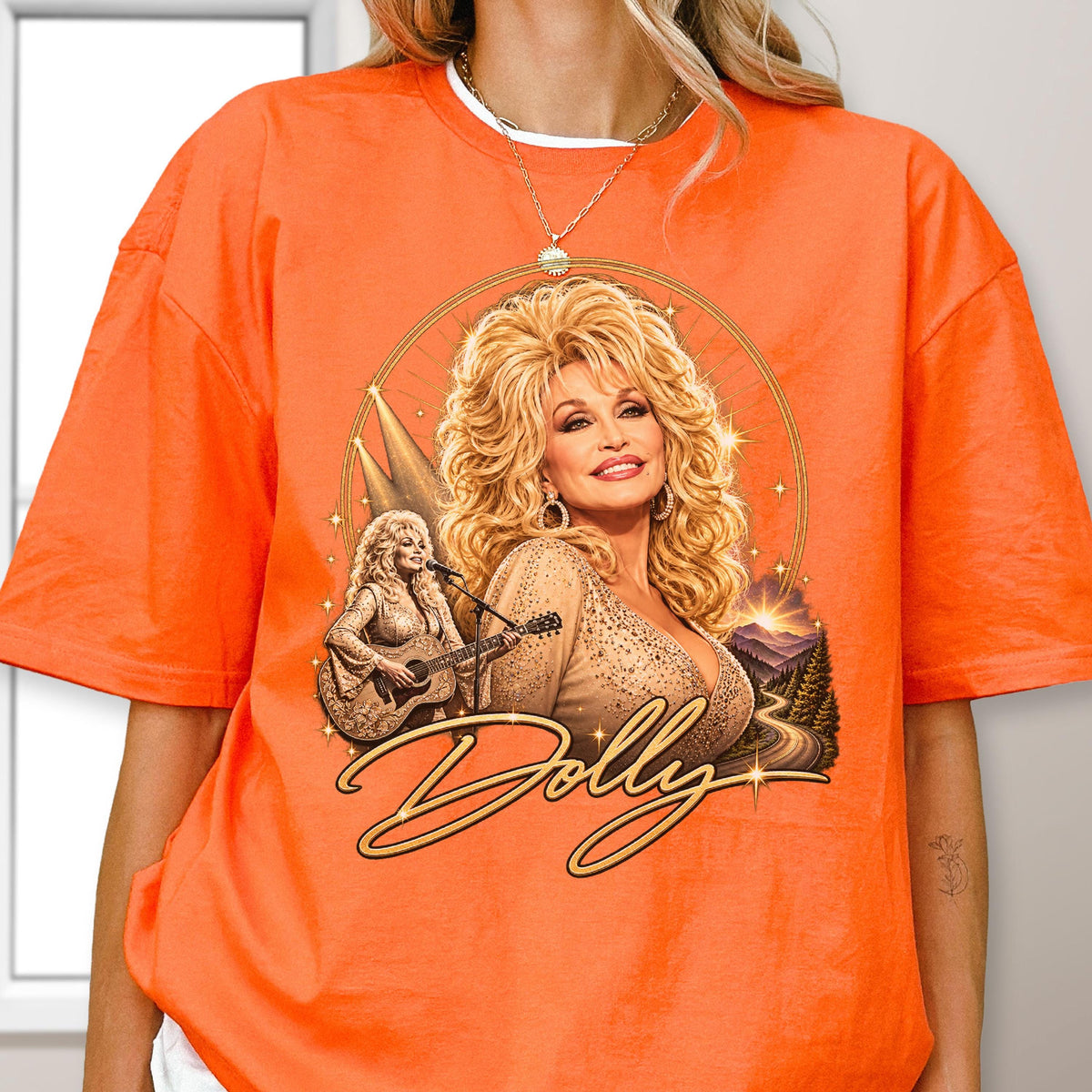 May include: An orange short-sleeve t-shirt featuring a graphic print of Dolly Parton with voluminous blonde hair, wearing a tan top. The design includes a smaller image of her playing an acoustic guitar, a mountain landscape, and the word Dolly.