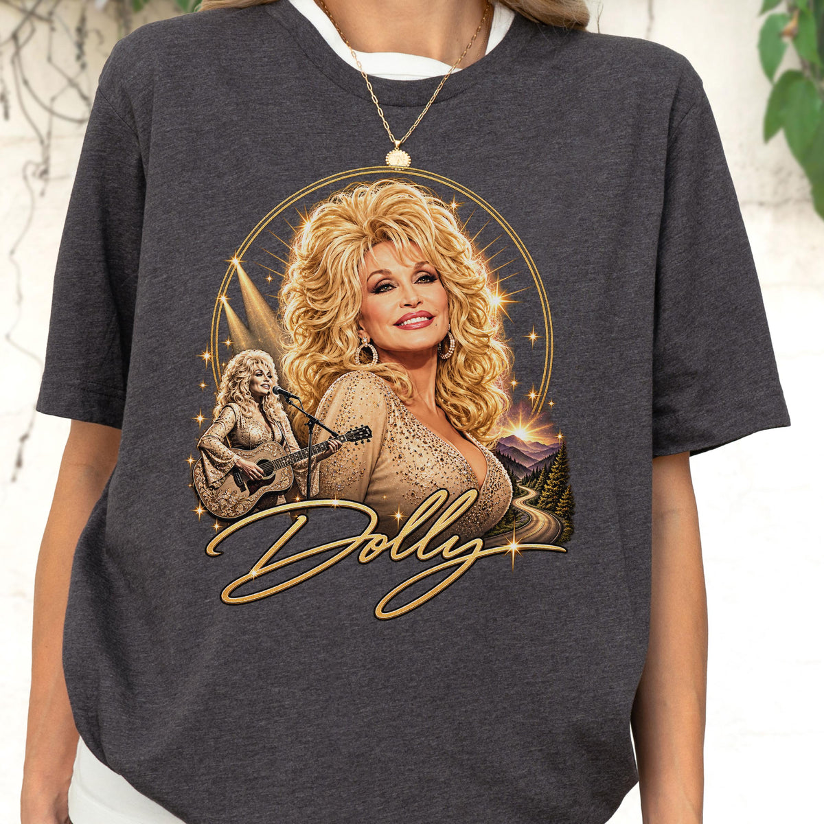 May include: A charcoal gray short-sleeve t-shirt featuring a graphic print of a blonde woman with voluminous hair. The design includes a smaller image of the woman playing an acoustic guitar, a mountain landscape, and the script text Dolly in gold.