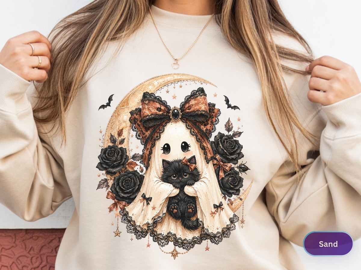 May include: A sand-colored crewneck sweatshirt featuring a graphic print of a ghost wearing a large lace bow and holding a black kitten. The design includes a crescent moon, black roses, small bats, and hanging star charms in shades of black and gold.