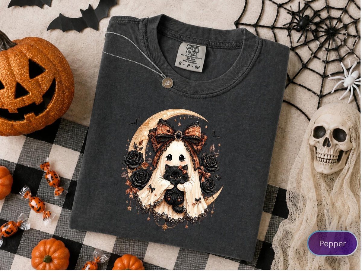 May include: A charcoal gray t-shirt featuring a graphic of a ghost wearing a large patterned bow and holding a black kitten. The design includes a crescent moon, black roses, and small stars. The shirt has a Comfort Colors brand tag at the neck.
