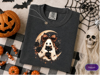 May include: A charcoal gray t-shirt featuring a graphic of a ghost wearing a large patterned bow and holding a black kitten. The design includes a crescent moon, black roses, and small stars. The shirt has a Comfort Colors brand tag at the neck.