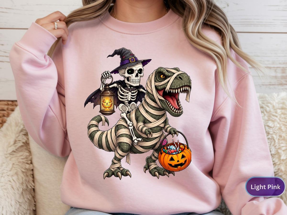 May include: A light pink crewneck sweatshirt featuring a graphic of a skeleton wearing a witch hat and cape, riding a T-Rex wrapped in mummy bandages. The skeleton holds a glowing jack-o'-lantern lantern, and the T-Rex carries a pumpkin candy bucket.