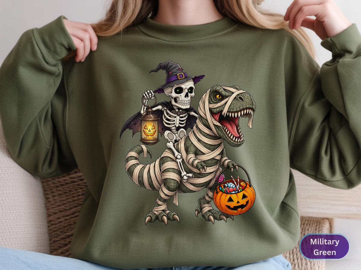 May include: A military green crewneck sweatshirt featuring a graphic of a skeleton wearing a purple witch hat and cape, holding a glowing jack-o'-lantern lantern while riding a mummified Tyrannosaurus Rex that carries an orange pumpkin candy bucket.