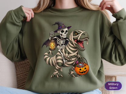 May include: A military green crewneck sweatshirt featuring a graphic of a skeleton wearing a purple witch hat and cape, holding a glowing jack-o'-lantern lantern while riding a mummified Tyrannosaurus Rex that carries an orange pumpkin candy bucket.