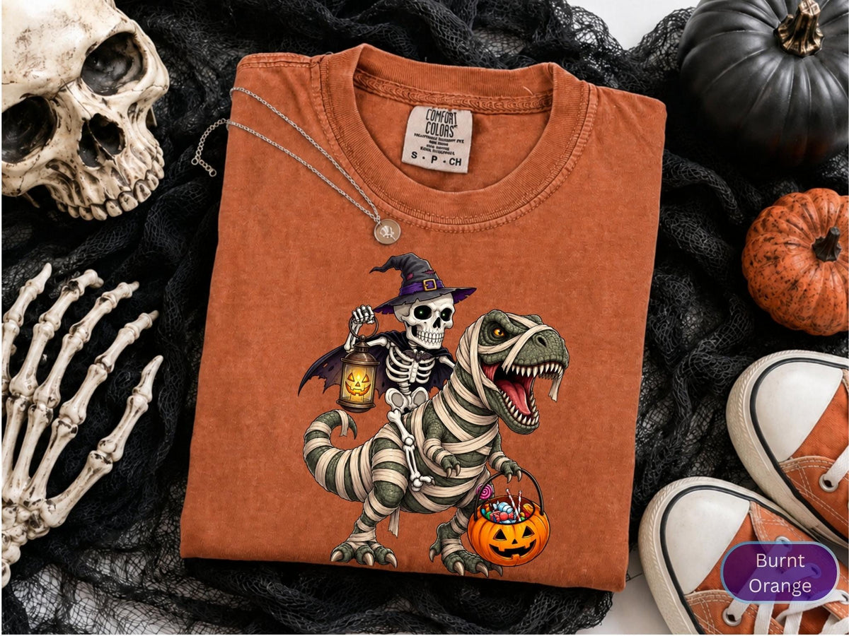 May include: A burnt orange t-shirt featuring a graphic of a skeleton wearing a purple witch hat and cape, riding a mummy-wrapped dinosaur. The skeleton holds a glowing jack-o'-lantern lantern, and the dinosaur carries a pumpkin candy bucket.
