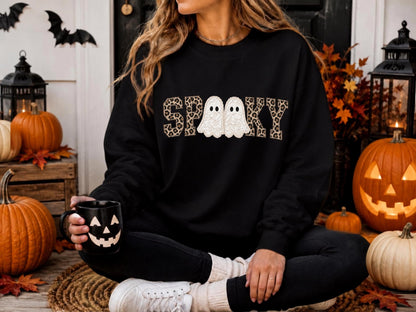May include: A black long-sleeve sweatshirt featuring the word SPOOKY printed in a brown and black leopard print pattern, with two white ghost illustrations replacing the letter O. The sweatshirt has a crew neckline and ribbed cuffs.