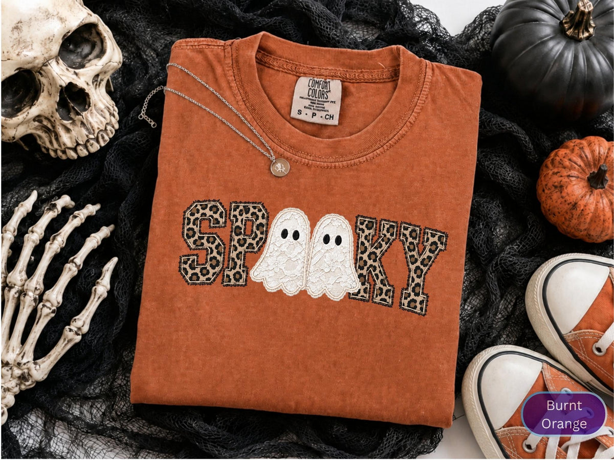 May include: A burnt orange t-shirt featuring the text SPOOKY in a leopard print pattern, with two white lace ghosts replacing the letter O. The shirt has a crew neckline and a Comfort Colors brand label on the inside collar.