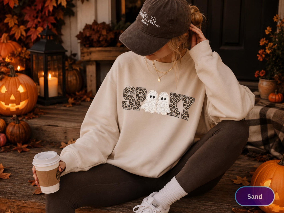 May include: A beige crewneck sweatshirt featuring the word SPOOKY in a black and grey leopard print pattern, with two white ghost illustrations replacing the letter O. The garment has long sleeves and a relaxed fit, worn with dark leggings.